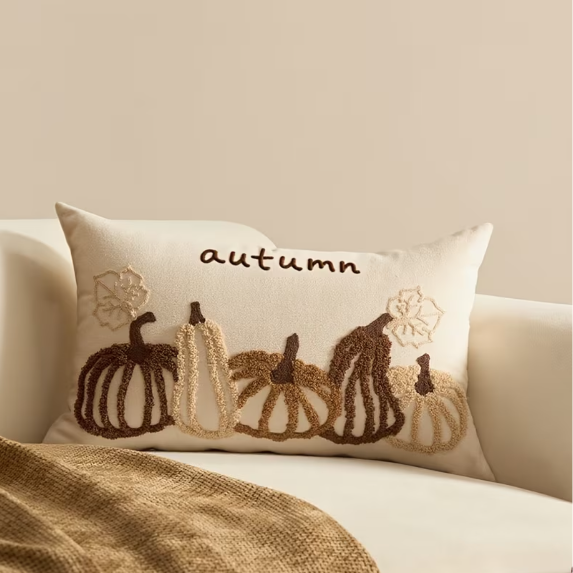 An autumn-themed throw pillow with embroidered pumpkins and the word 'autumn' in beige and brown, resting on a cream sofa with a brown blanket.